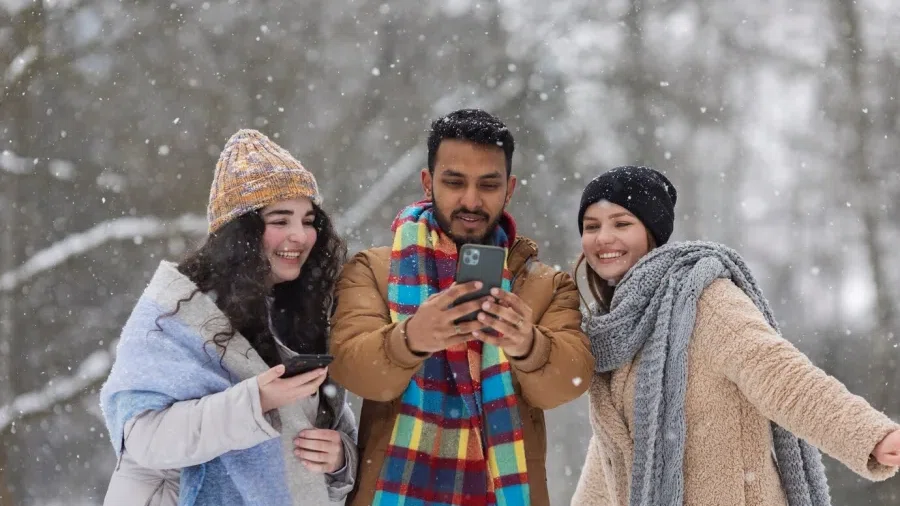 3 Menschen schauen auf ein Handy und sehen fröhlich aus. Sie stehen in einem Wald und es schneit.