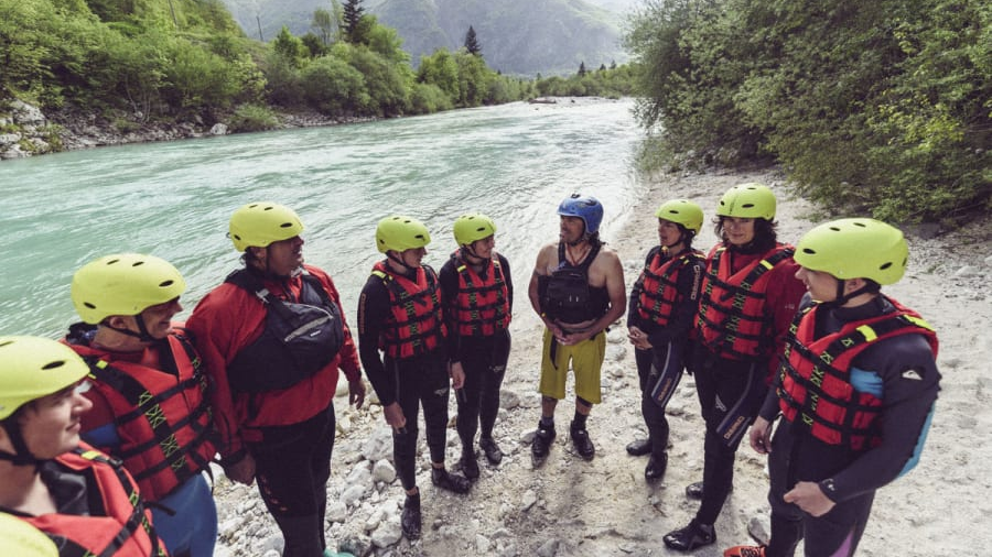 Mehrere Menschen stehen in der Natur vor einem Fluss. Ausgerüstet sind sie mit Helmen und Schwimmwesten. Ein Mann erklärt den anderen etwas.