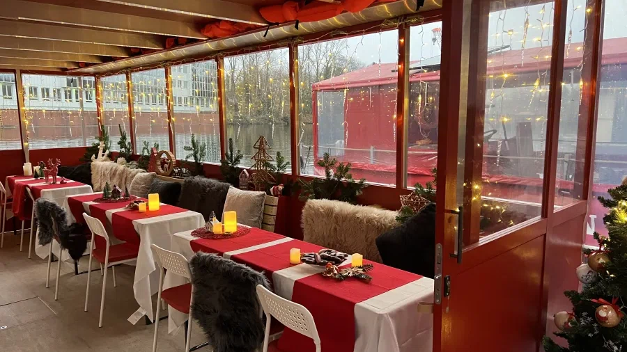 Stühle und Tische mit weihnachtlichem Gedeck stehen in einem Raum auf einem Boot mit einer Fensterwand, die mit Lichterketten geschmückt ist.