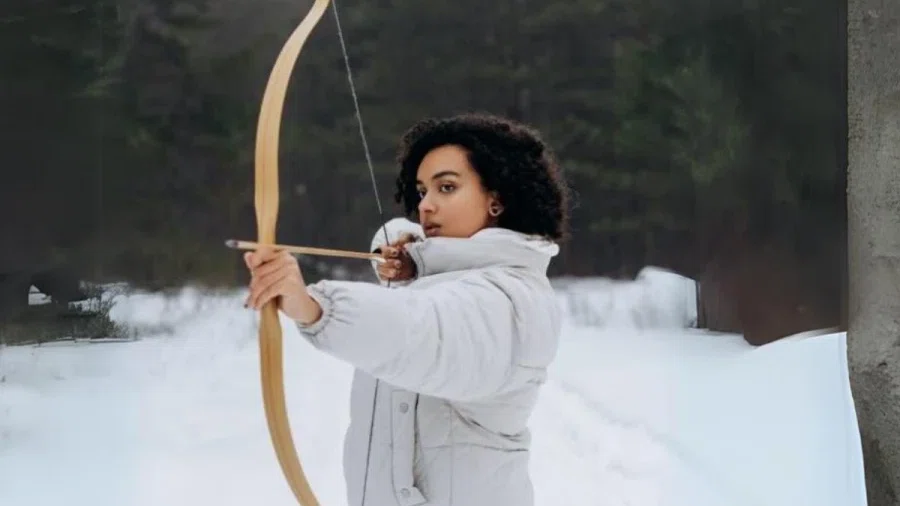 Eine Frau steht in einem Wald mit Schnee. Sie trägt Winterbekleidung und eine Pfeil und Bogen in der Hand. Den Pfeil spannt sie im Bogen.