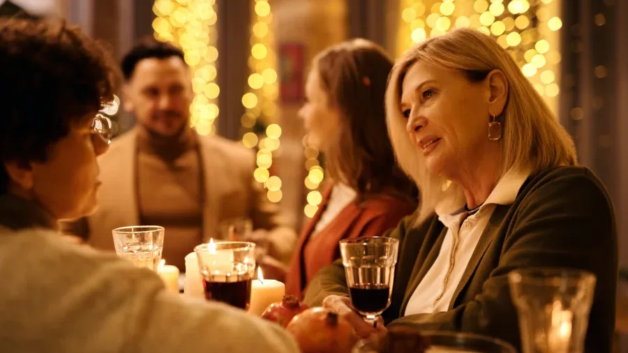 4 Menschen sitzen mit einem Weinglas in der Hand an einem Tisch und unterhalten sich beim Dinner. Im Hintergrund sind Lichterketten die leuchten.